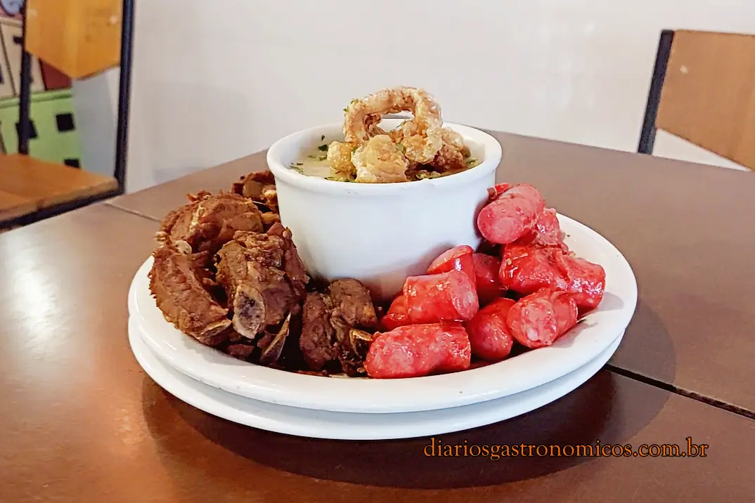 Porção de angu com queijo, com o torresmo por cima, junto com costelinha de porco e linguiça no Bar da Gema, na Tijuca, Rio de Janeiro. Plate on a wooden table with a white cup of creamy soup topped by a crispy crackling ring, surrounded by brown fried pork rib and red sausage chunks on a white platter.