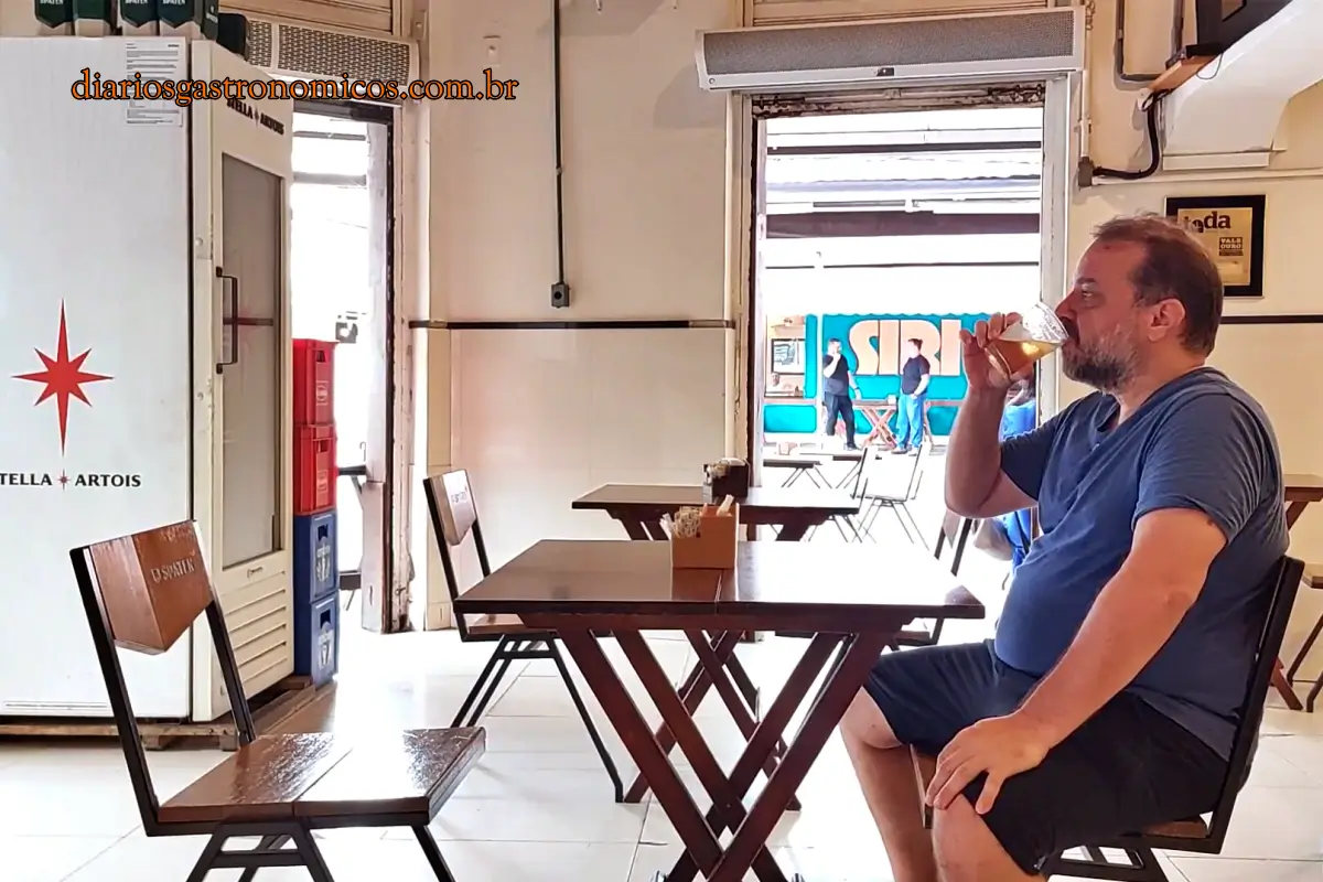 O salão interno no Bar da Gema, típico boteco carioca no bairro da Tijuca. Man in a blue shirt and black shorts sits at a wooden table inside a bright cafe, drinking a glass of beer to the side.