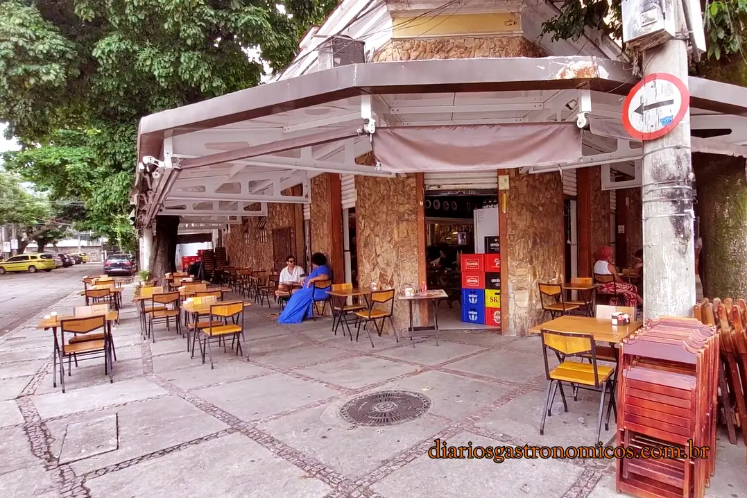 Bar da Gema, boteco que fica numa esquina do bairro da Tijuca, no Rio de Janeiro, com mesas ao ar livre. Outdoor cafe on a sidewalk with wooden tables and yellow chairs in front of a stone-faced building.