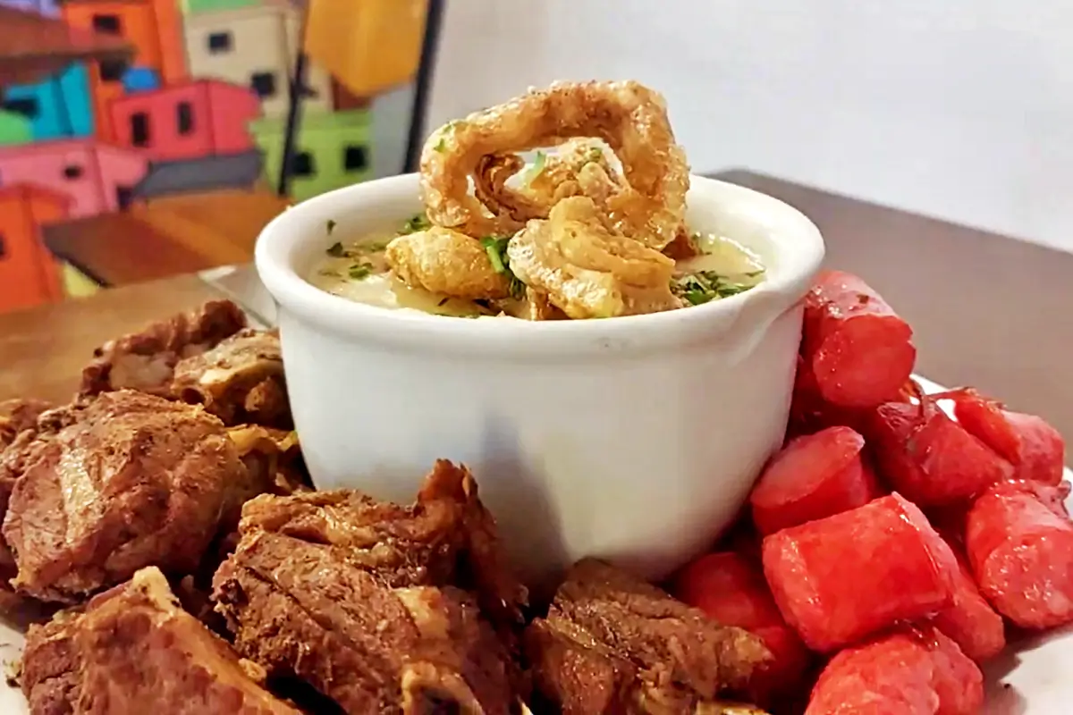 Angu com torresmo, costelinha de porco e linguiça no Bar da Gema na Tijuca. Creamy soup in a white bowl topped with crispy crackling, with braised pork rib and red salsages cubes on the plate nearby.