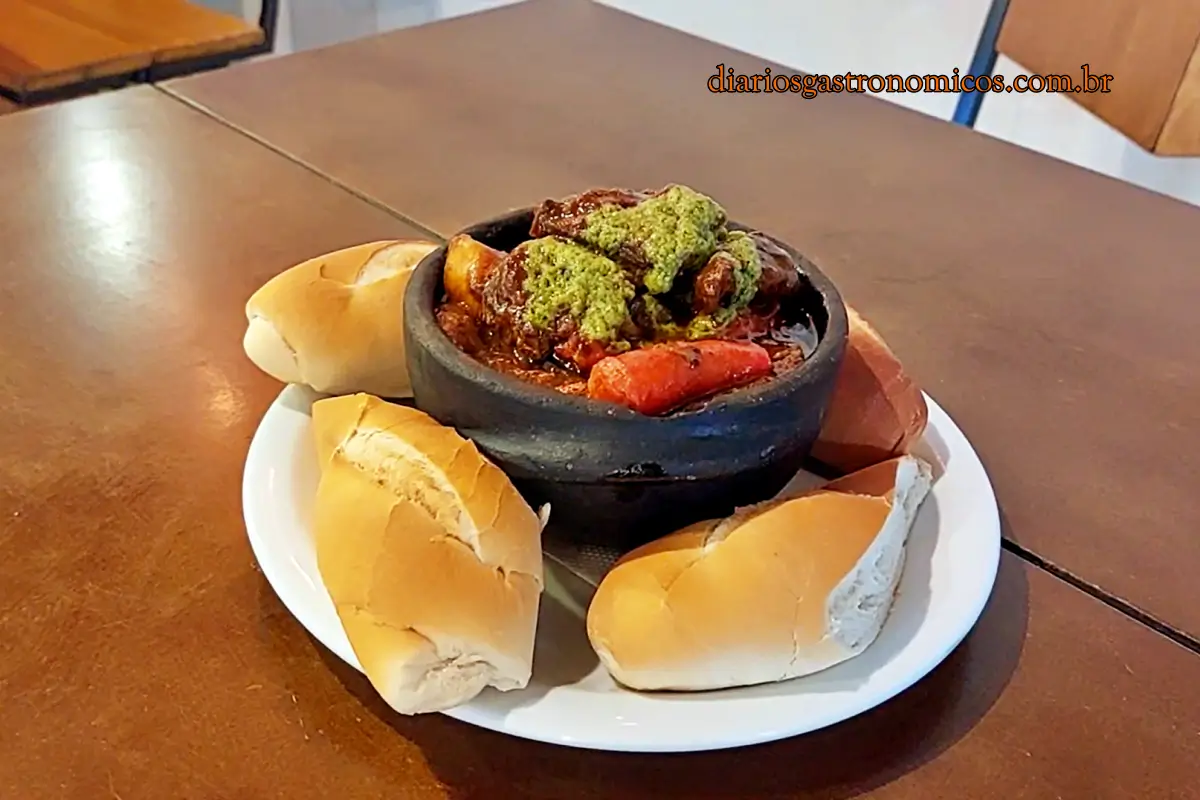Ensopado de peito bovino com linguiça no Bar da Gema, na Tijuca. Braised meat stew with green herb sauce in a black stone pot, surrounded by crusty bread on a white plate.
