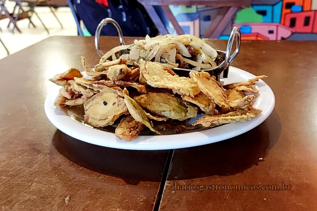 Porção de jiló frito tipico de boteco brasileiro no Bar da Gema na Tijuca. Plate of crispy fried scarlet eggplant with onion garnish on a white platter, set on a wooden table in a casual restaurant.
