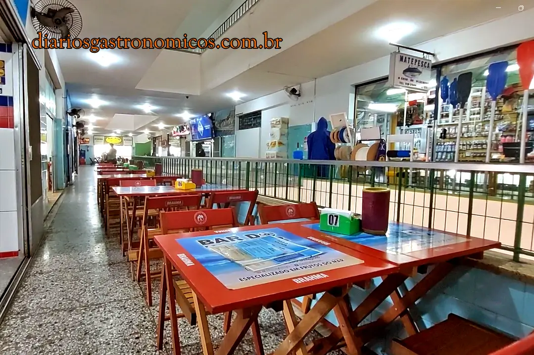 Mesas do Bar do Wagner, no segundo andar do Mercado de Peixe de Niterói, Rio de Janeiro Market hall with red tables and wooden chairs lined along a railing; table 07 visible and a row of shops on the right; watermark 'diariosgastronômicos.com.br' in the corner.