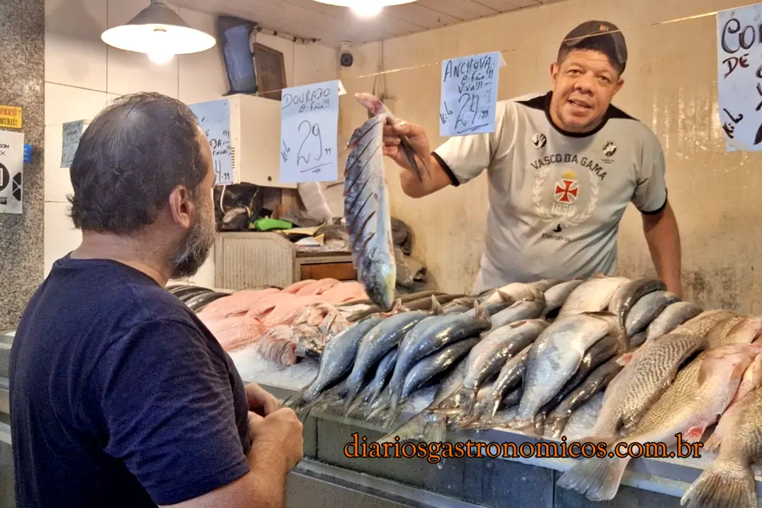 Vendedor exibe um peixe na Peixaria do Guimarães, no Mercado de Peixe de Niterói, Mercado de São Pedro, Fishmonger at a market holds a large fish up for inspection while a customer watches; rows of fish on ice behind them.