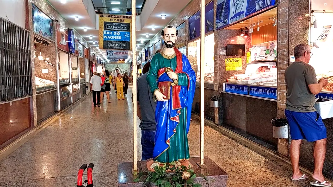 Imagem de São Pedro no Mercado de Peixe de Niterói, Mercado de São Pedro, Statue of a bearded saint in a green and blue robe holding a book, standing on a pedestal in a busy indoor market corridor with shopfronts on both sides and shoppers in the background,