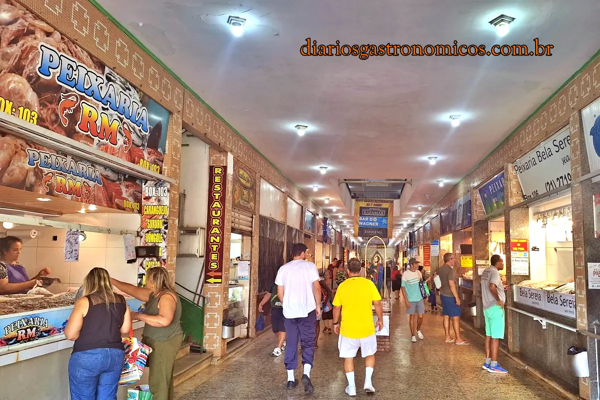 Movimento nas peixarias do corredor principal do Mercado de Peixe de Niterói, Indoor market corridor with seafood stalls on the left and diners, shoppers walking along tiled walkway under bright ceiling lights.