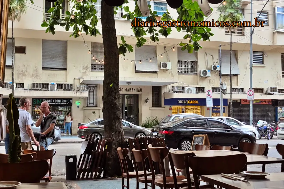 Imagem da entrada do bar, na Rua Bento Lisboa, no catete. Demi Boteco, boteco de comida francesa no Largo do Machado, no Rio de Janeiro. Outdoor sidewalk cafe with wooden chairs and tables, string lights, and a tree in the foreground overlooking a city street.
