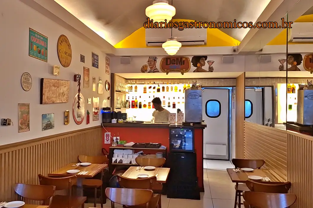 O ambiente do Demi Boteco, boteco de comida francesa no Largo do Machado, no Rio de Janeiro. Café/bar interior with wooden tables and chairs, wall decor, and a backlit counter displaying bottles behind a staff member.6d13a1