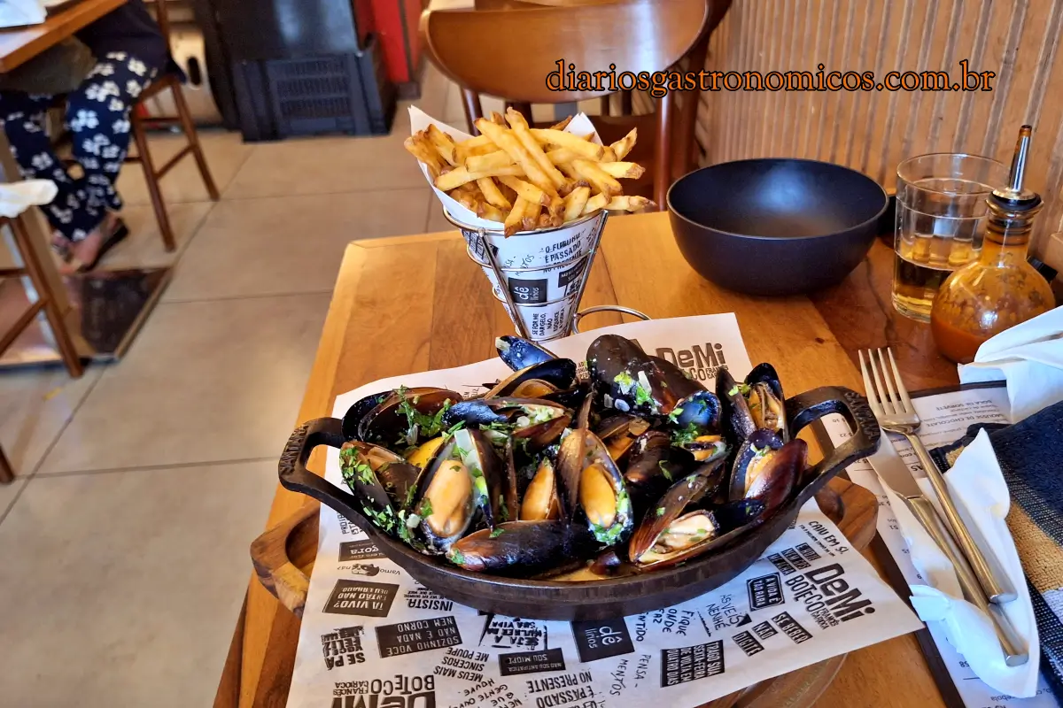 Porção de Moules Frites do Demi Boteco, boteco de comida francesa no Catete, no Rio de Janeiro. Cast-iron skillet of mussels in herb sauce on a wooden table, with a paper-cone of fries nearby.