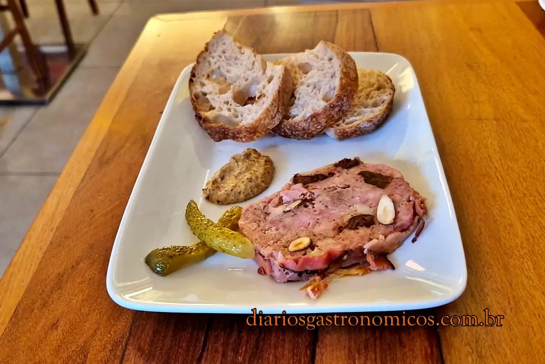 patê de campagne do Demi Boteco, boteco de comida francesa no Largo do Machado, no Rio de Janeiro. É priduzido pela marca carioca Cochon Rouge. Plate with sliced rustic bread, a pink beef roast with almonds, and a dollop of mustard; pickles on the side; watermark at bottom indicating diariosgastronomicos.com.br