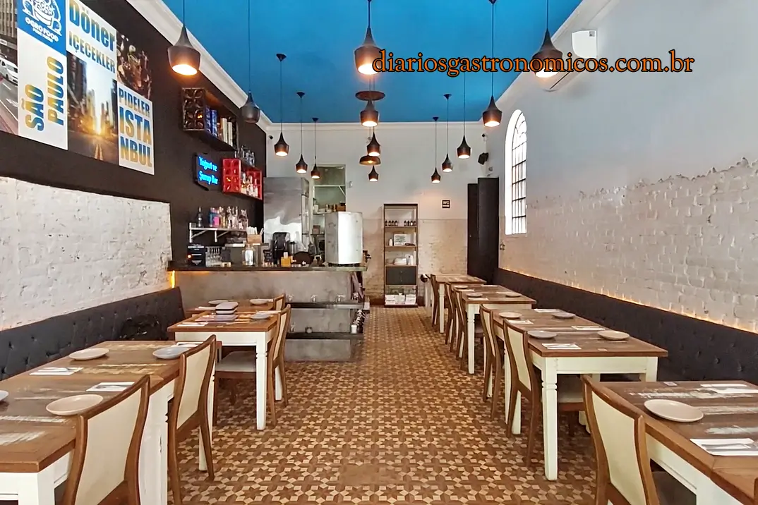 Restaurante Kebab Salonu, de estilo casual e intimista, no bairro de Perdizes, em São Paulo. Bright casual restaurant interior with a blue ceiling, pendant lights, white brick walls, and rows of wooden tables and chairs. Open-kitchen cafe feel with tiled floor and posters on the left wall, seen from the dining area.