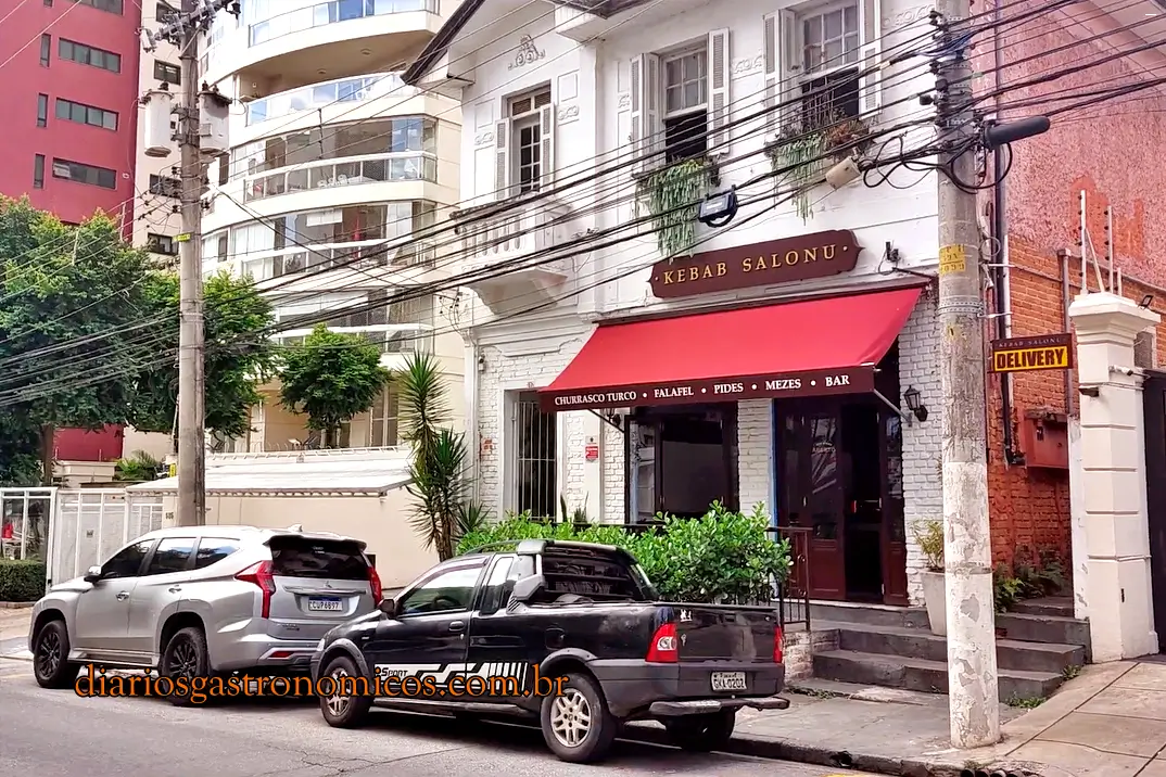 O restaurante Kebab Salonu que fica numa casa histórica no bairro de Perdizes, em São Paulo White brick storefront with a red canopy reading 'KEBAB SALONU' on a city street; two cars parked along the curb in front.