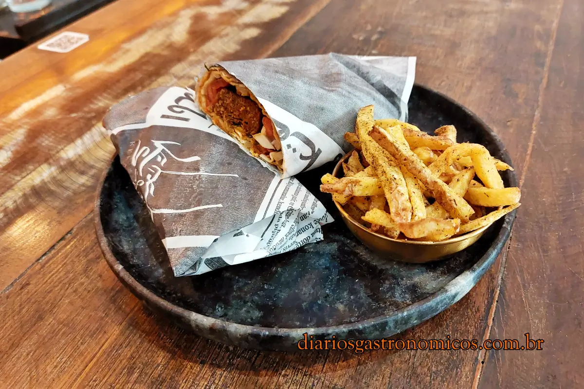 Lahmacum Kofte, comida turca, culinária turca. Wrap-style sandwich opened to show filling, served with seasoned fries on a dark plate on a wooden table, with diario's watermark.