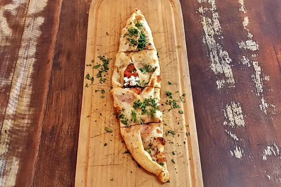A Pide turca, famosa por seu formato de barco, Flatbread on a wooden board topped with melted cheese, chopped herbs, and greens, sliced to reveal tomato inside.