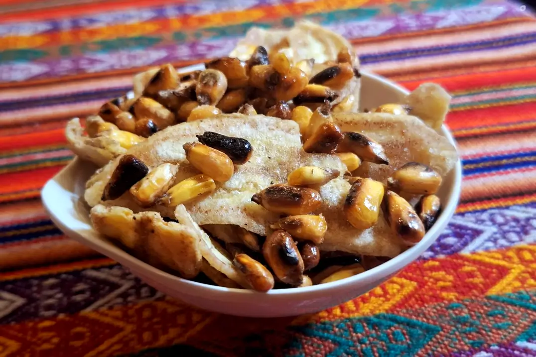 Chifles e cancha servidos no restaurante peruano Doña Bertha, nos Campos Elíseos, em São Paulo. White bowl of fried plantain chips topped with roasted corn kernels on a colorful woven tablecloth.