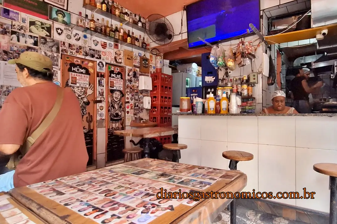 Interior do Bar do Adalto na Glória com mesas compartilhadas, balcão e paredes decoradas com fotos de clientes e lembranças da boemia carioca.