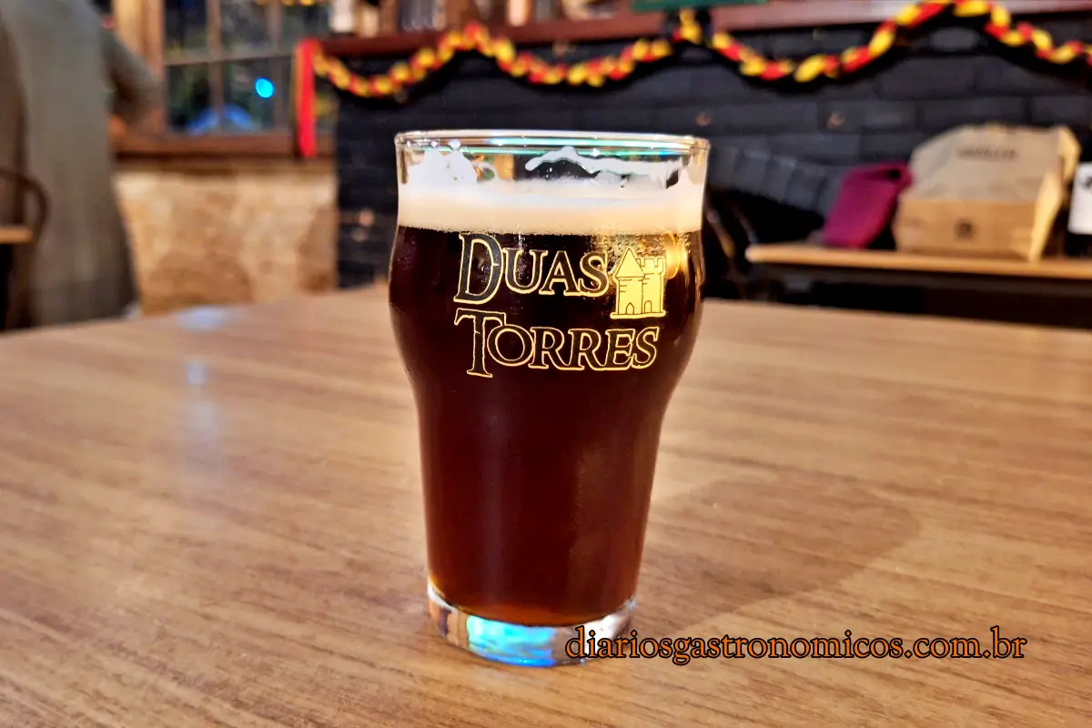 Copo de cerveja artesanal âmbar da Cervejaria Duas Torres sobre mesa de madeira, com o salão temático ao fundo em Petrópolis.