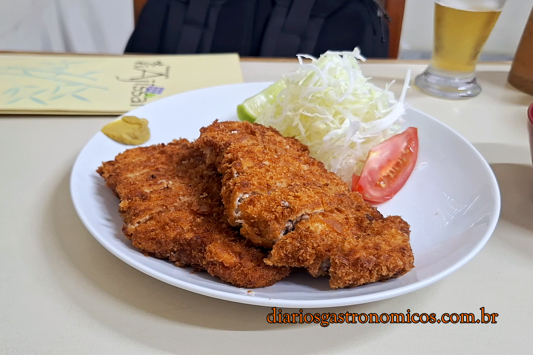 Prato de tonkatsu servido no Ajissai, com duas fatias de lombo suíno empanadas em panko, acompanhadas de repolho fatiado, tomate e limão.